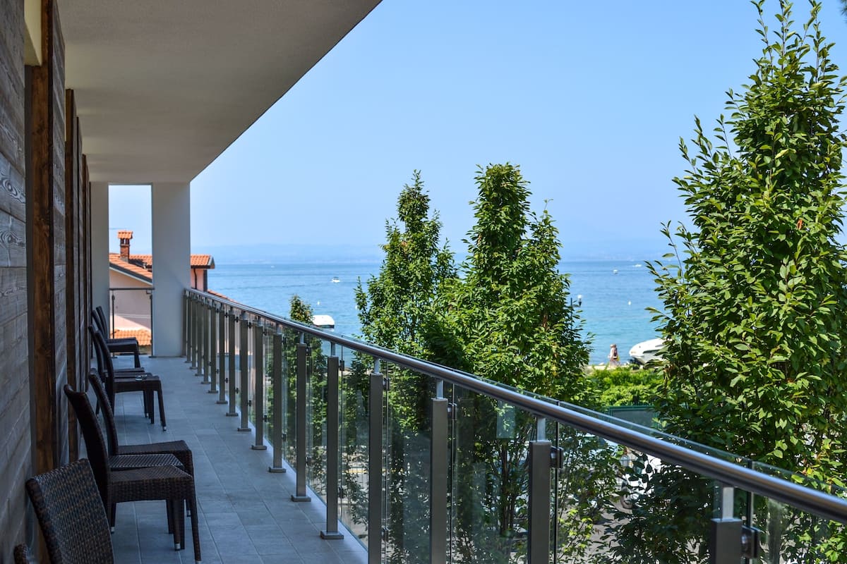 A balcony with wicker chairs, a glass railing, and a view of the sea and distant buildings.