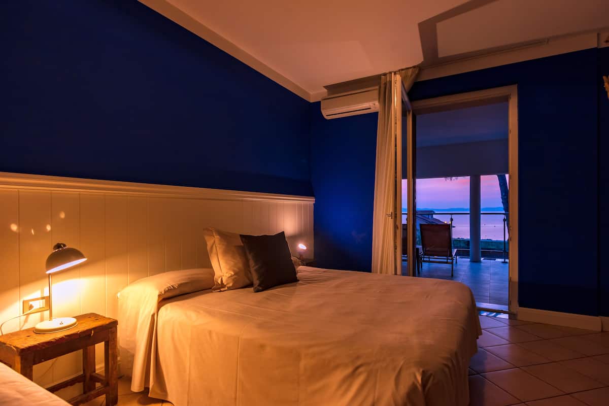 A hotel room with a bed, bedside table with a lamp, and a view of the beach through an open door.
