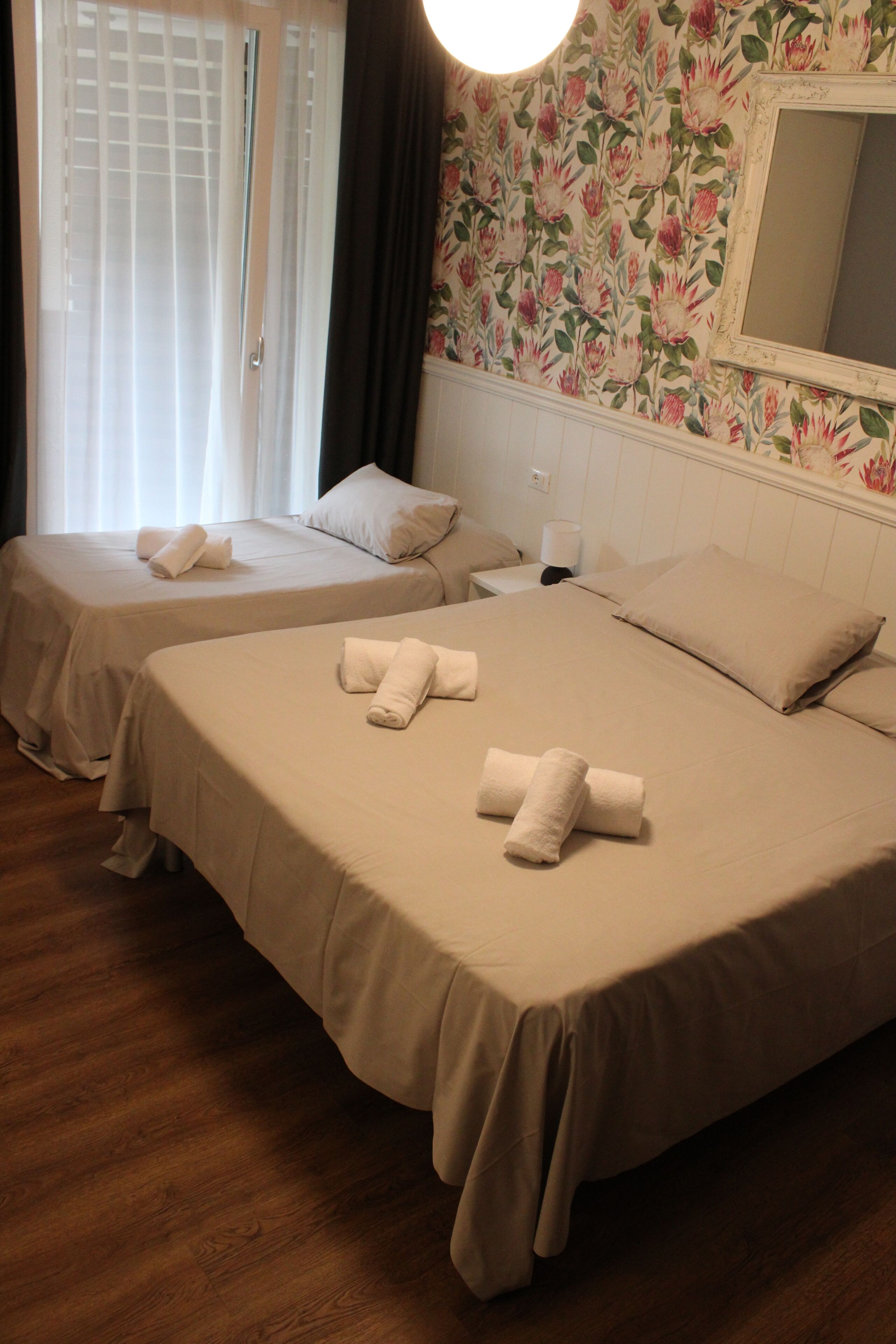 Two single beds with towels, a wooden floor, a floral wallpaper, and a window with curtains.