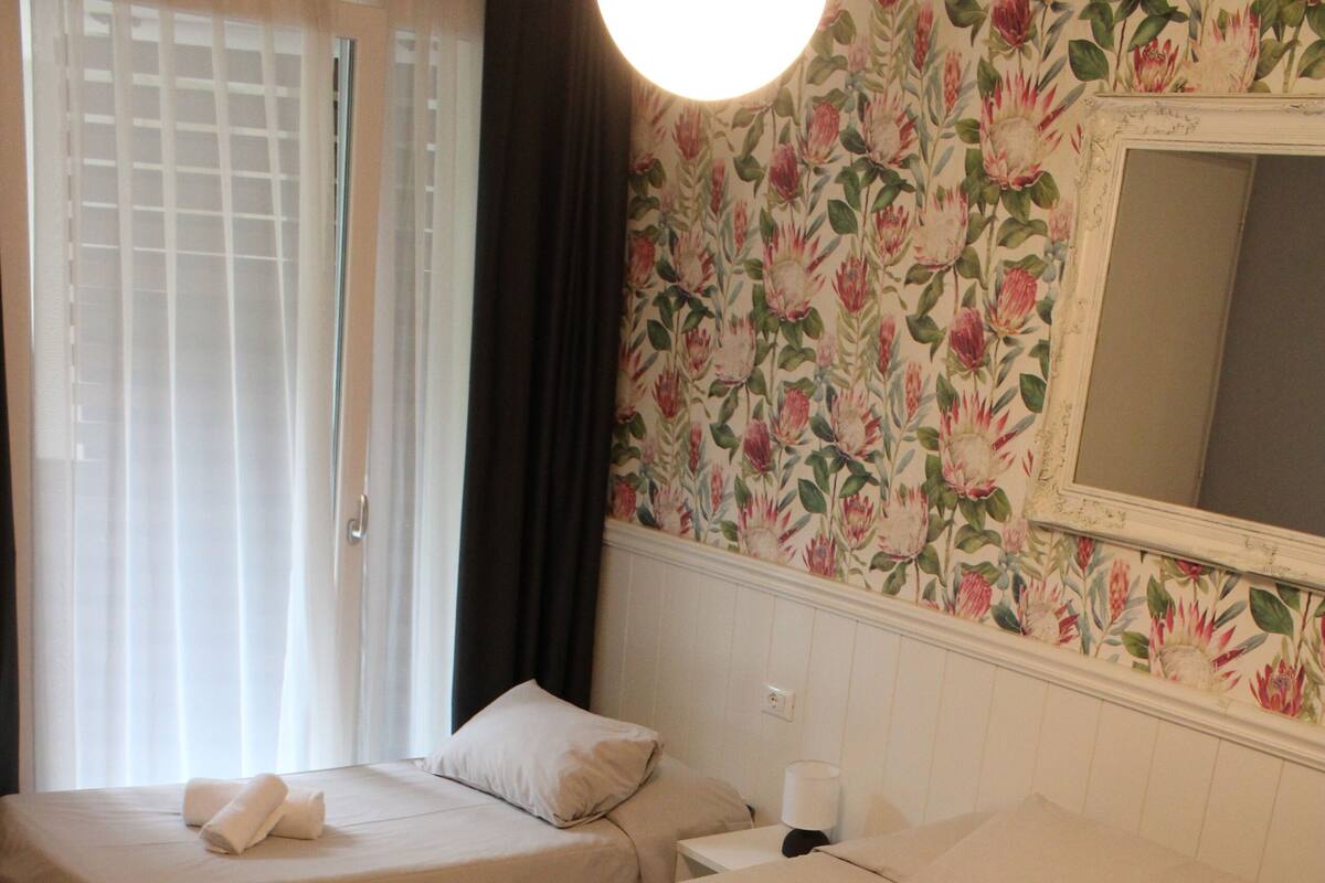 Two single beds with towels, a wooden floor, a floral wallpaper, and a mirror on the wall.
