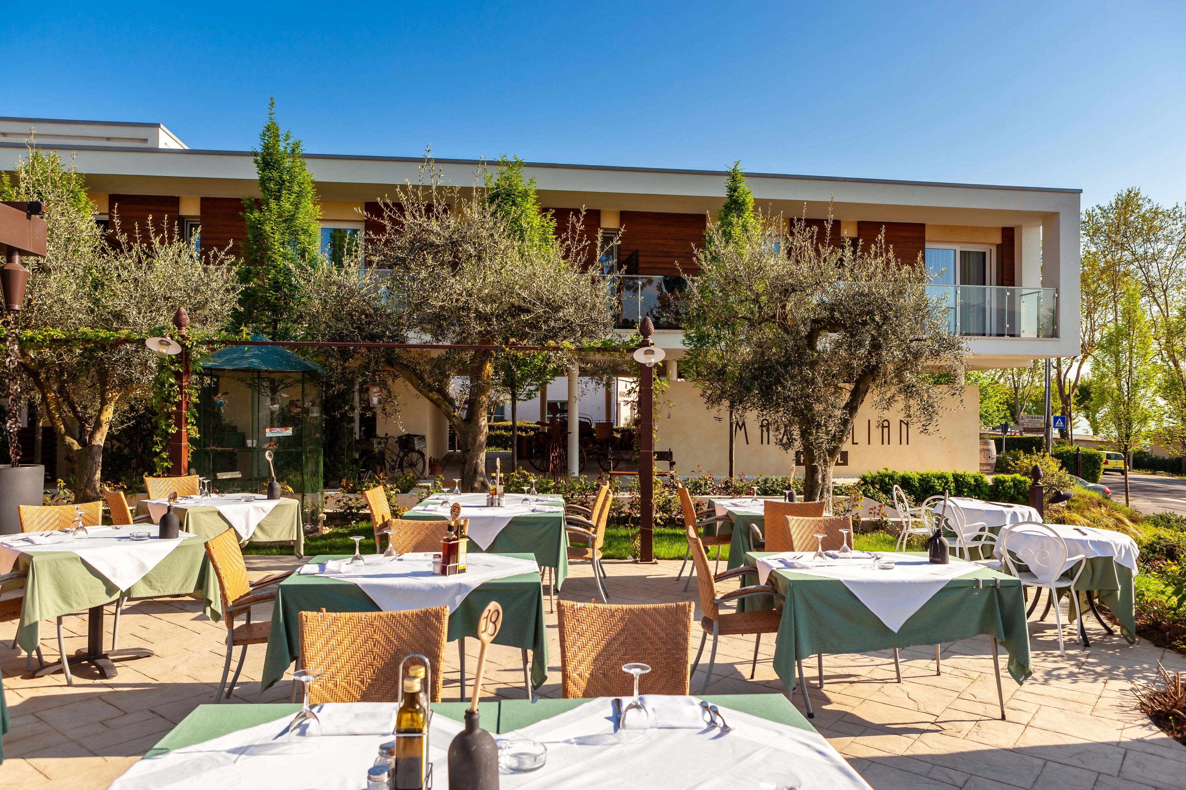 Un espace extérieur aménagé pour les repas, avec des tables et des chaises, un bâtiment doté d’un balcon et des arbres en arrière-plan.