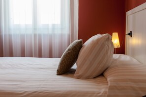 A neatly made bed with striped bedding and two pillows, a bedside lamp, and a window with sheer curtains.