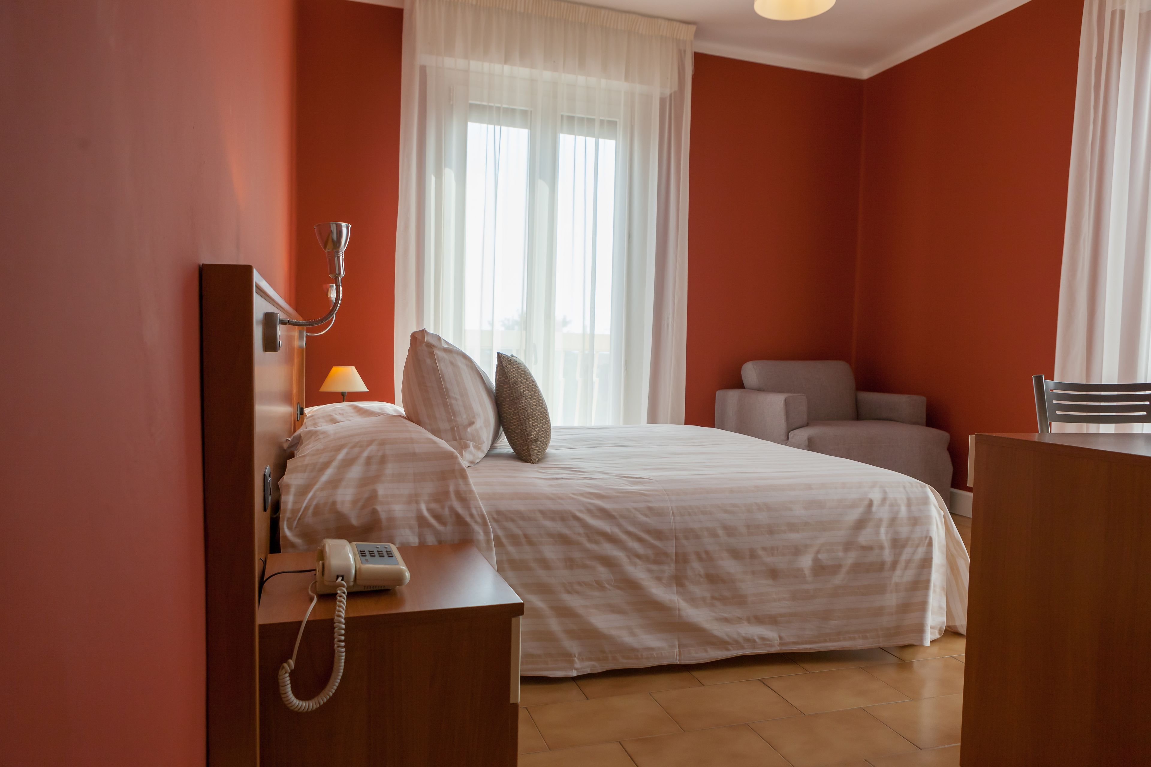 A hotel room with a bed, bedside table with a telephone, a chair, and a desk with a lamp.
