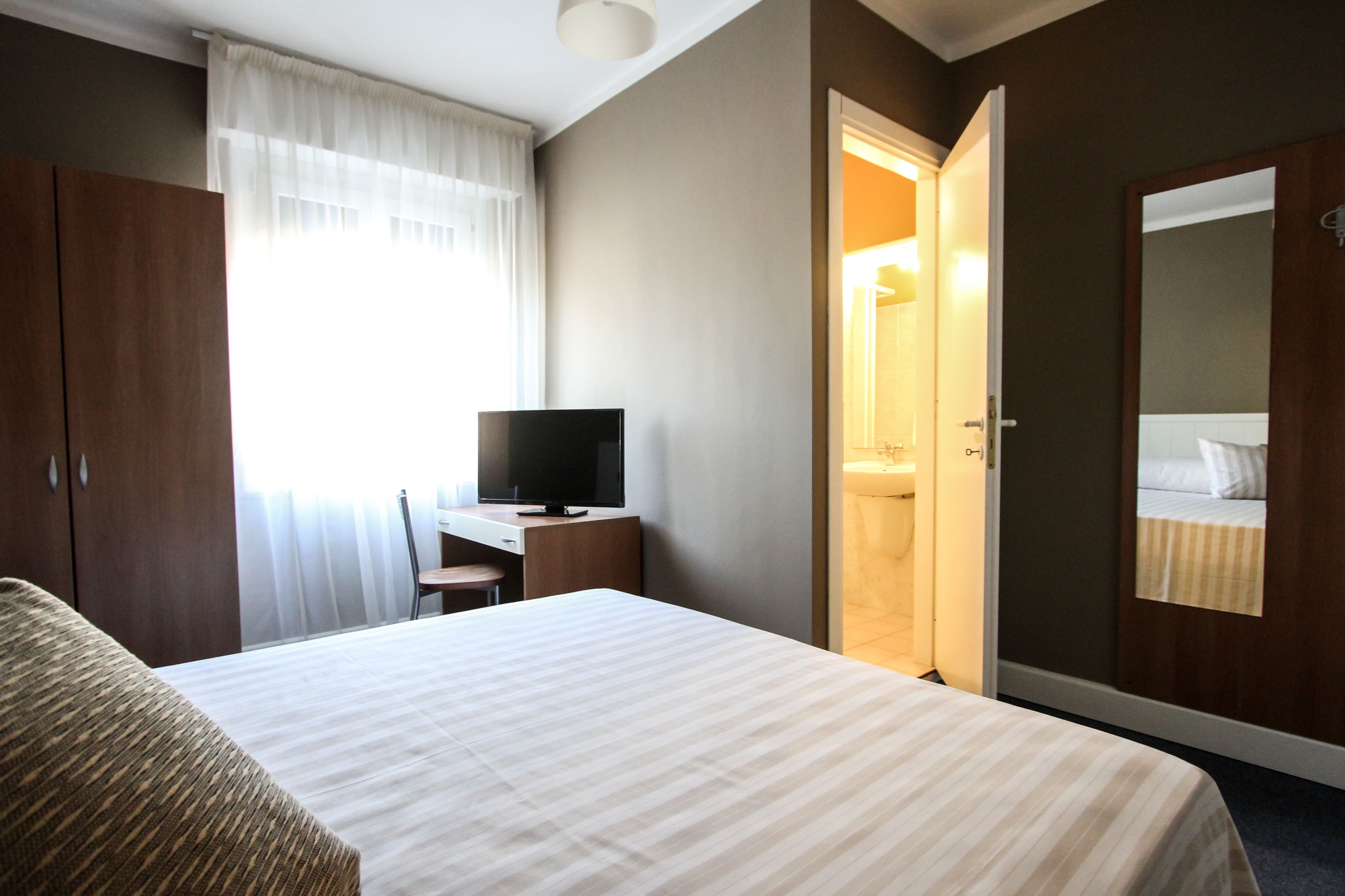 Camera d'albergo con un letto, una scrivania con televisore, un armadio e un bagno visibile attraverso una porta aperta.