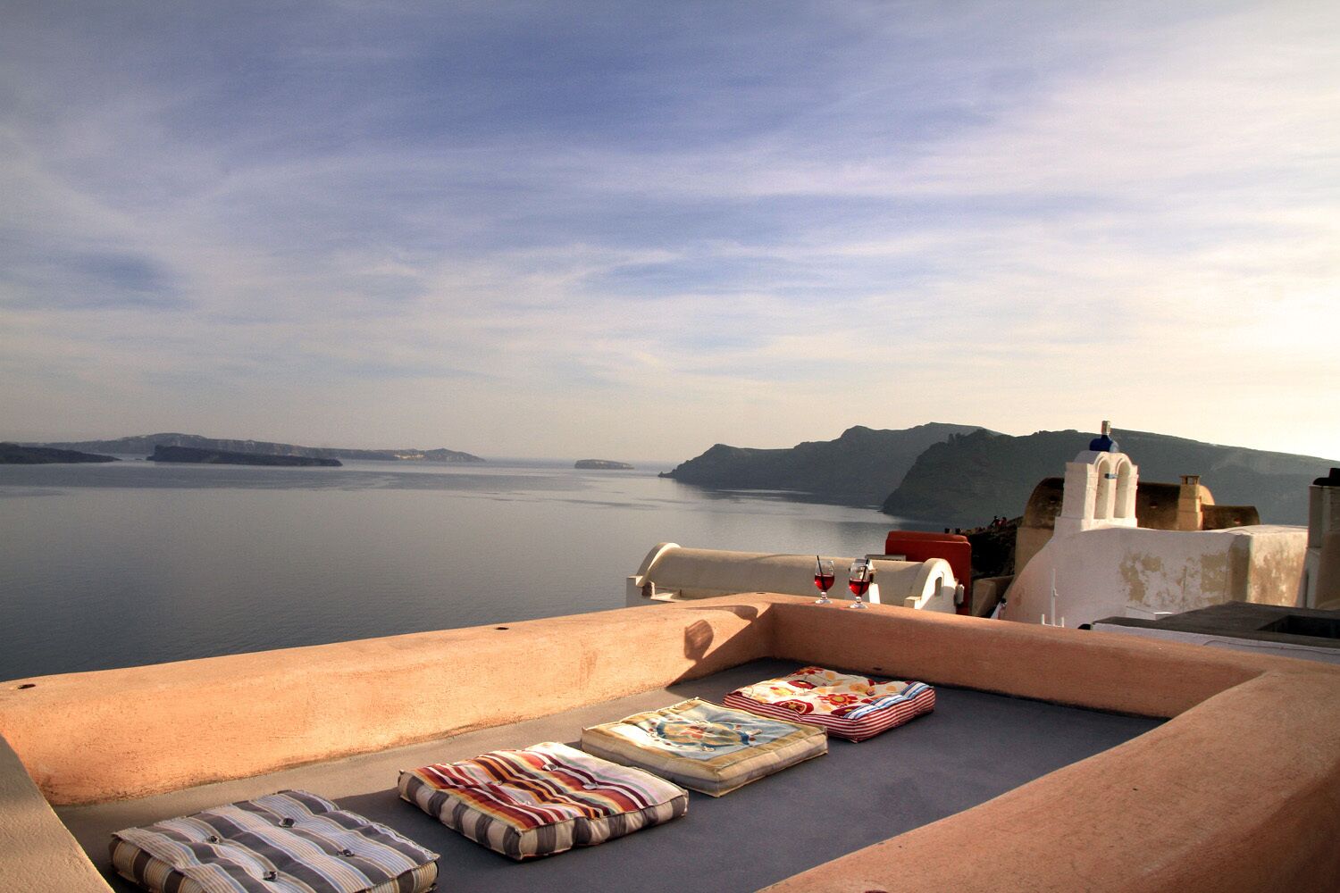 Une terrasse offrant une vue sur la mer et les îles lointaines, avec un coin salon agrémenté de coussins et une bouteille de vin rouge.