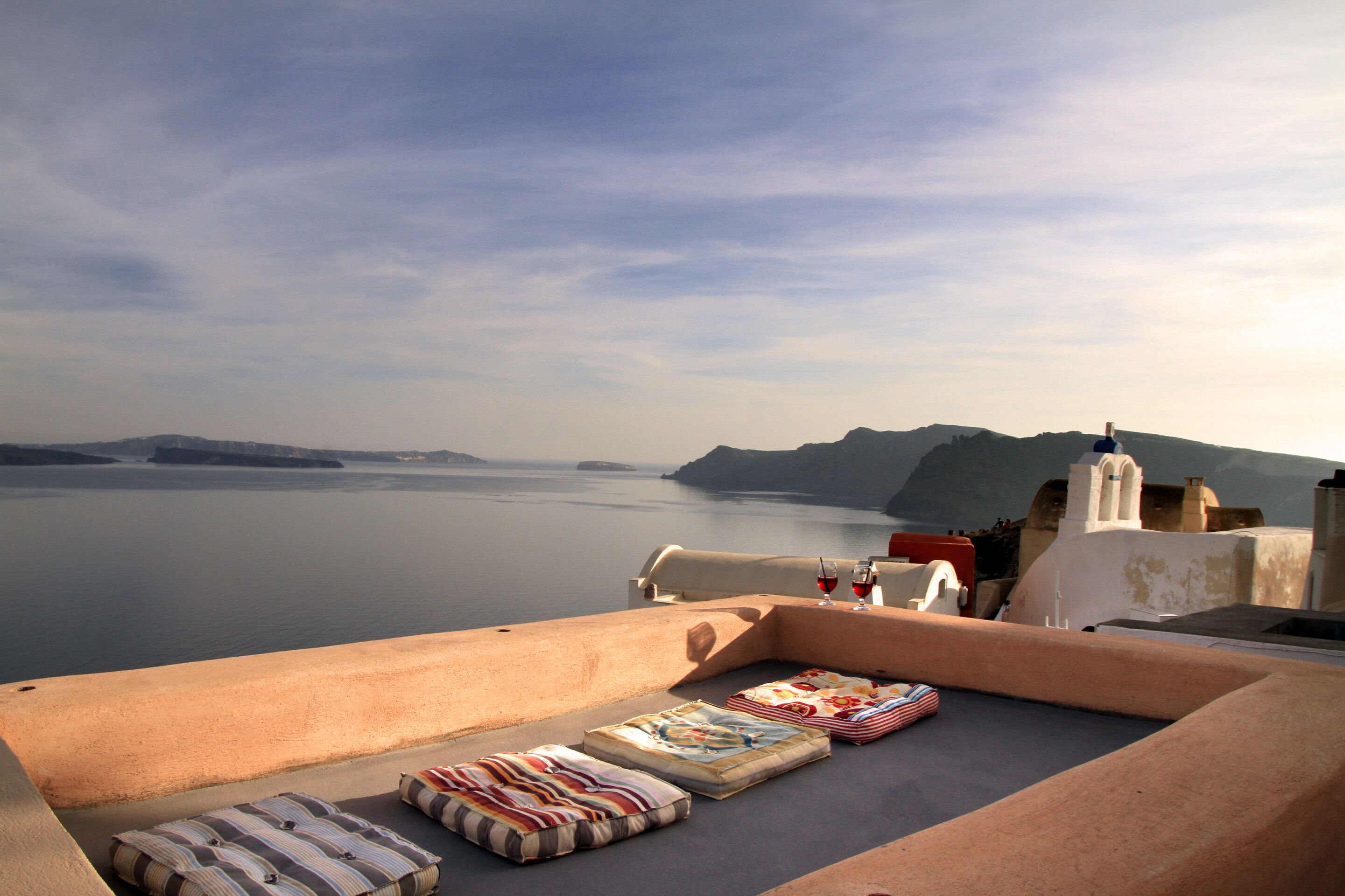 A terrace with a view of the sea and hills, featuring a table with a bottle and glasses, and several cushions.