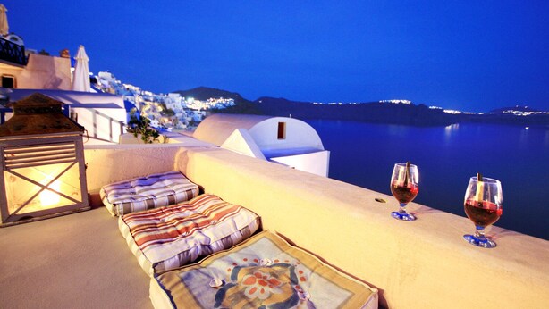 Terraza frente al mar con vistas a un pueblo costero de noche, con un sofá acolchado y dos copas de vino tinto.