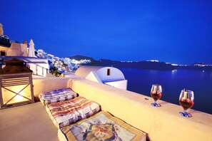 A seaside terrace with a view of a coastal town at night, featuring a sofa with cushions and two glasses of red wine. - Alafouzou Cave Loft (Santorini)