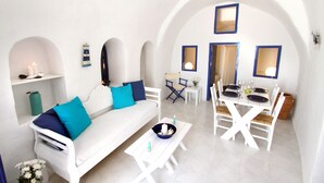 Una sala de estar blanca con sofá, mesa de centro y mesa de comedor. Hay cojines azules en el sofá y una mesita con un jarrón y velas. La habitación tiene paredes blancas y arcos.