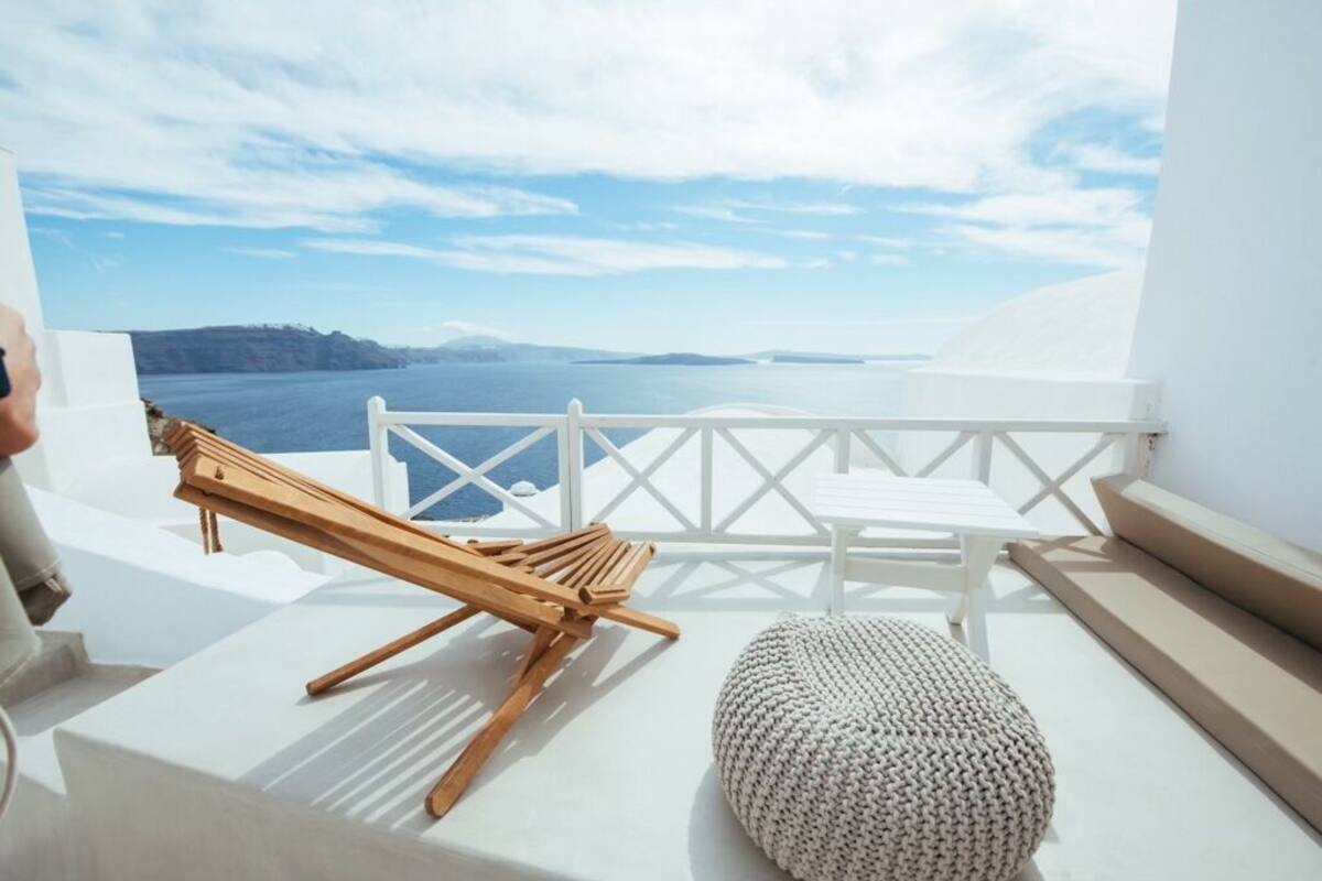 Un balcón con una silla de lounge de madera, un banco blanco acolchado y vista al mar y a la costa lejana.