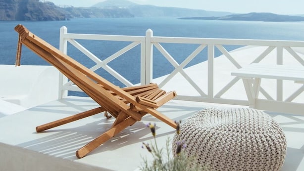 Una terraza blanca con una silla de lounge de madera y un otoman tejido, con vistas a un paisaje marino.