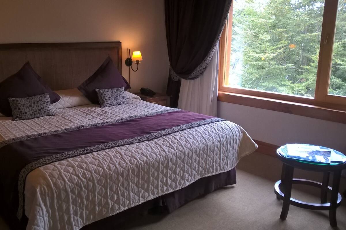 A neatly made bed with a headboard, two bedside lamps, and a window with curtains.