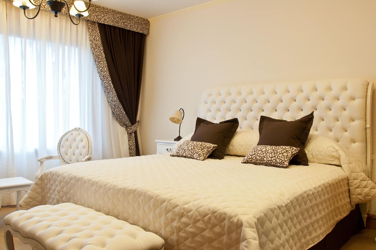 A large bed with a tufted headboard, two pillows, a bedside table, a lamp, a chair, and a bench.
