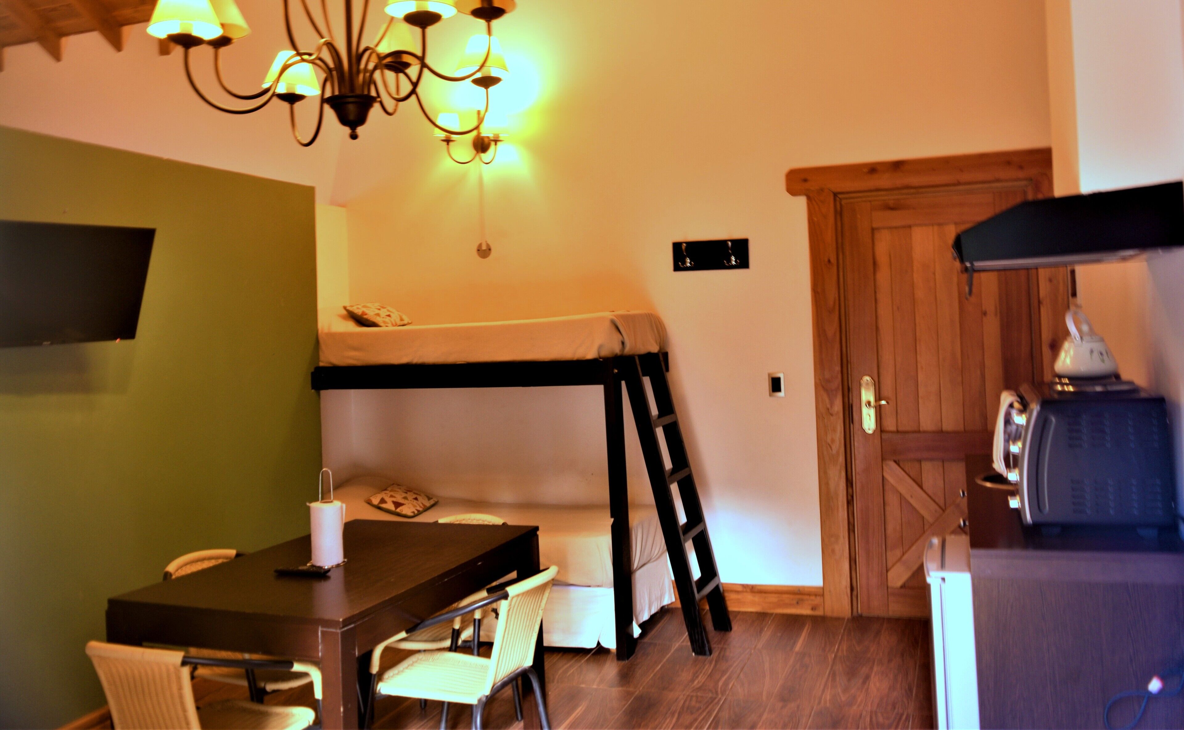 Una habitación compacta con una cama individual alta y otra inferior, una mesa de comedor con sillas, una araña y una puerta de madera.