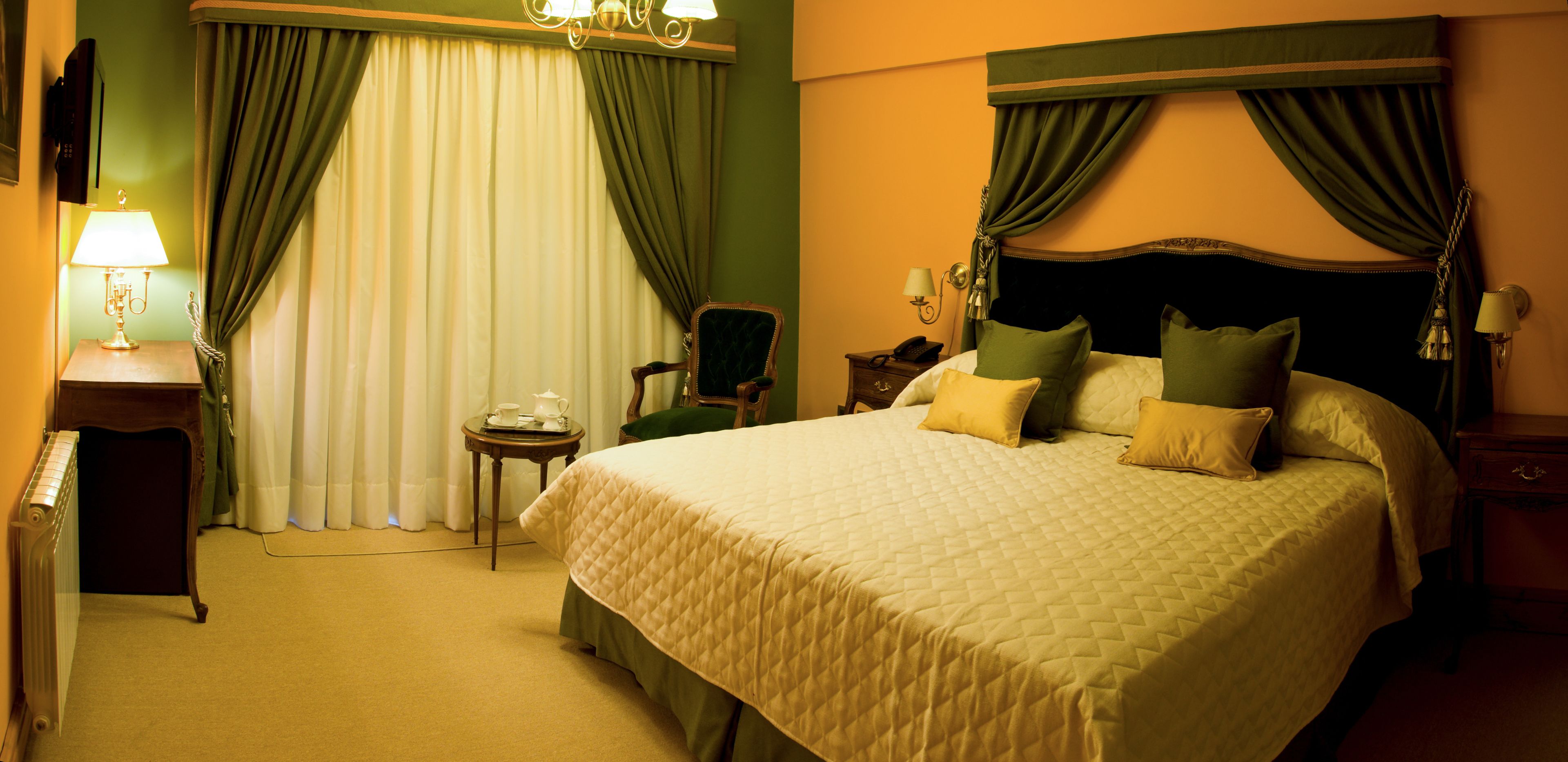 A hotel room with a large bed, green curtains, a small table with a lamp, and a chair.