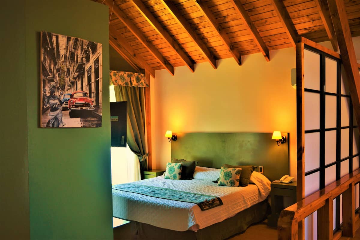 A bedroom with a bed, two bedside lamps, a nightstand, a window with curtains, and a wooden ceiling.