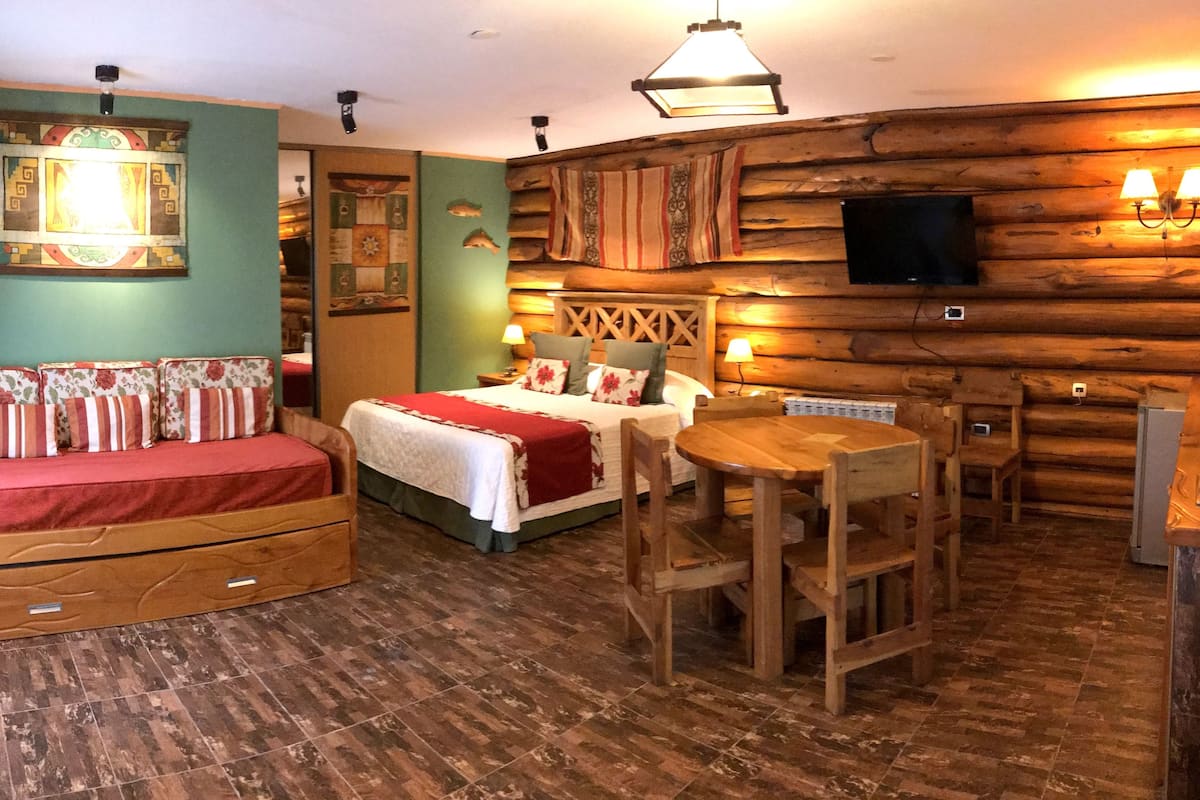 A cozy room with a bed, a sofa, a dining table, and a TV mounted on a wooden wall.