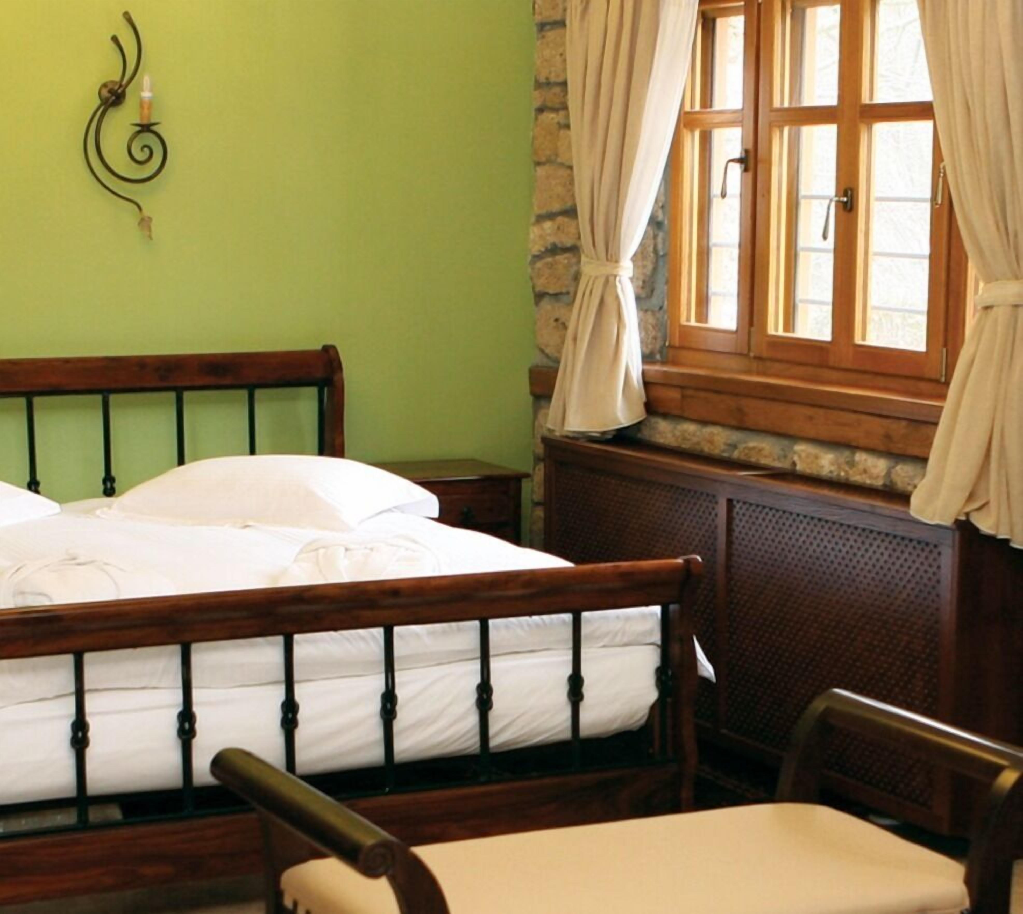 Un dormitorio con cama de madera, una silla, una mesita, una ventana con cortinas y una lámpara fijada a la pared.