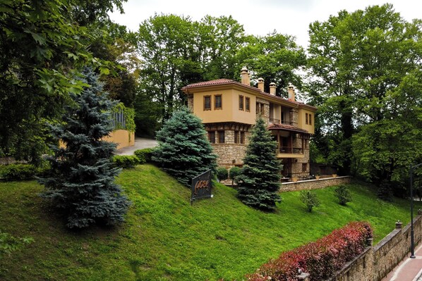 A two-story house with a stone facade and a red-tiled roof, surrounded by lush greenery and trees.