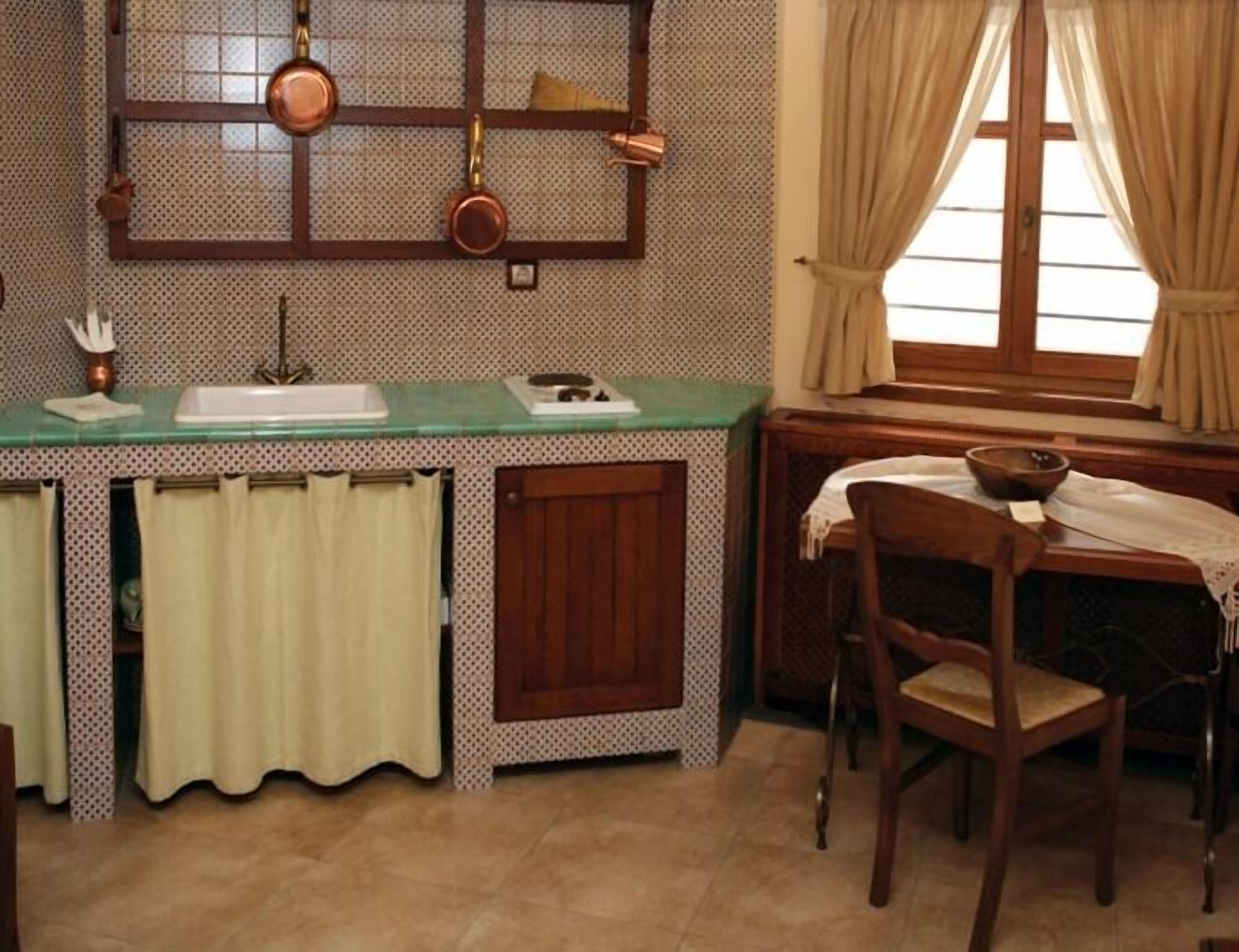 A kitchen with a green countertop, copper pots, a wooden cabinet, a window with curtains, and a wooden chair.