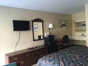 Una camera d'albergo con un letto, una cassettiera, una televisione, una scrivania con una lampada, uno specchio e un bagno con un lavandino e uno specchio.
