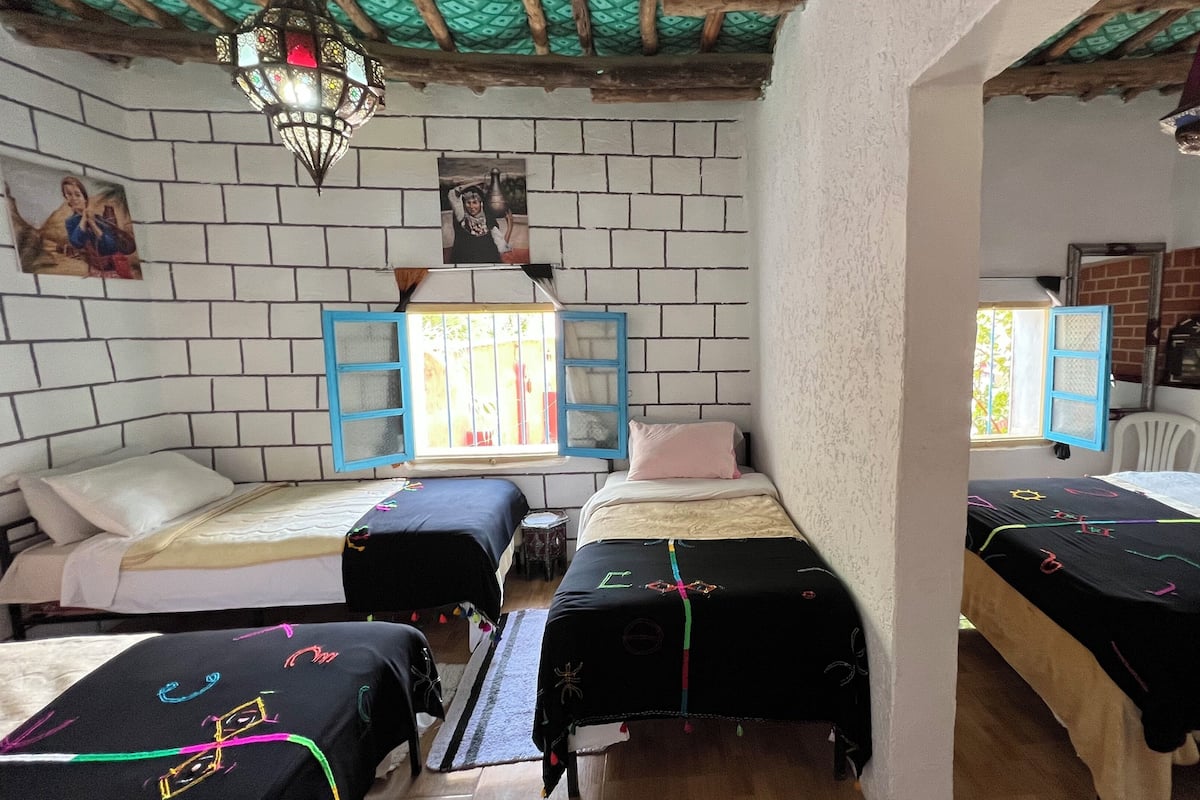 Um quarto com duas camas, uma janela com persianas azuis, um candeeiro suspenso e uma parede de tijolos.