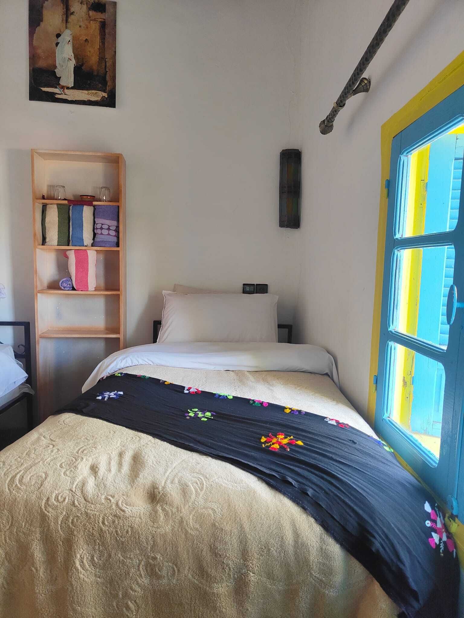 Quarto com cama, prateleira de madeira com toalhas dobradas, quadro na parede e janela com moldura azul.