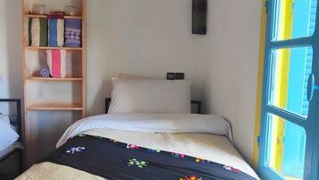 Quarto com cama, prateleira de madeira com toalhas dobradas, quadro na parede e janela com moldura azul.