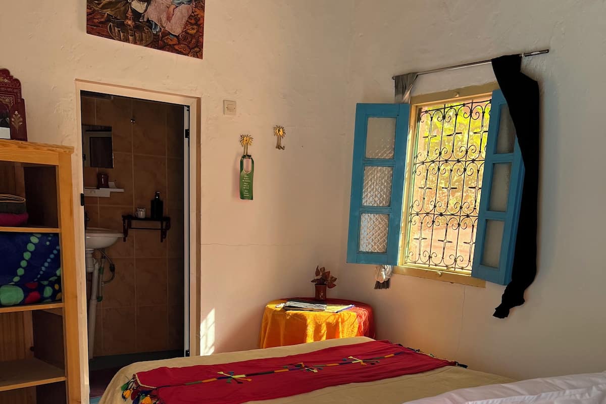 Quarto com cama, guarda-roupa de madeira, pequena mesa com toalha vermelha, janela com persianas azuis e um quadro na parede.