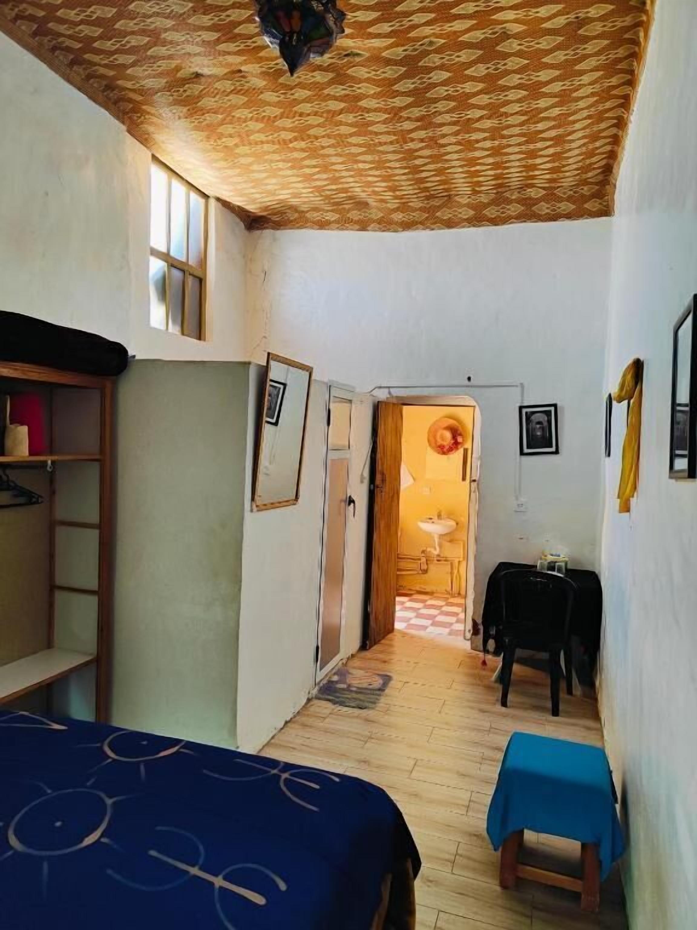 Ein schmaler Flur mit Holzbalkendecke, einem Bett mit blauer Decke, einem kleinen Tisch mit blauer Tischdecke und einem Kleiderschrank.