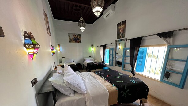 Habitación con dos camas, un ventanal con contraventanas azules, una lámpara colgante y un aire acondicionado de pared.