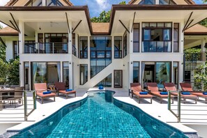 Una casa moderna de dos pisos con piscina, sillones de descanso y una escalera que sube al piso superior.