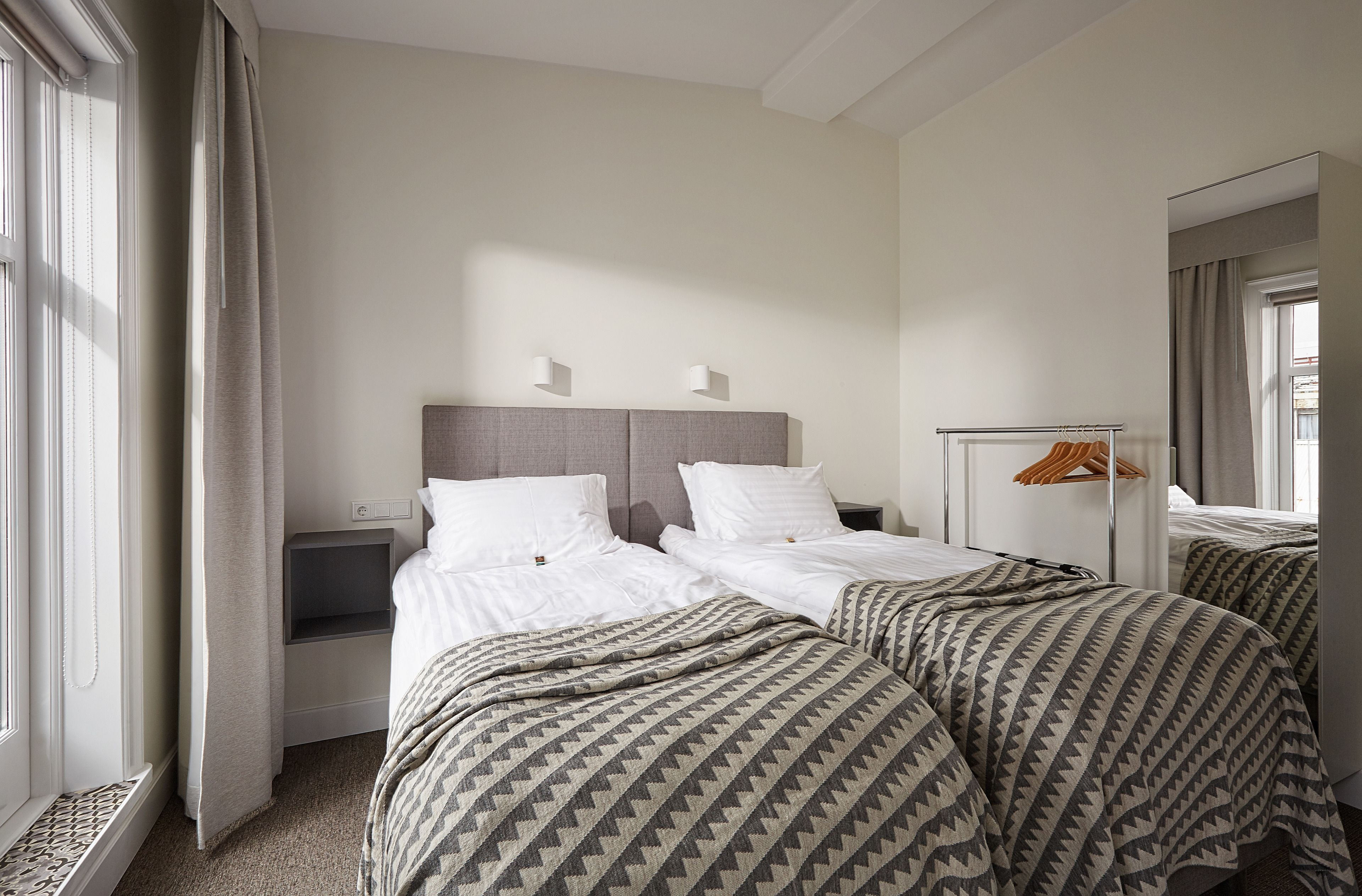 A hotel room with two beds, a bedside table, a mirror, and a window with curtains.
