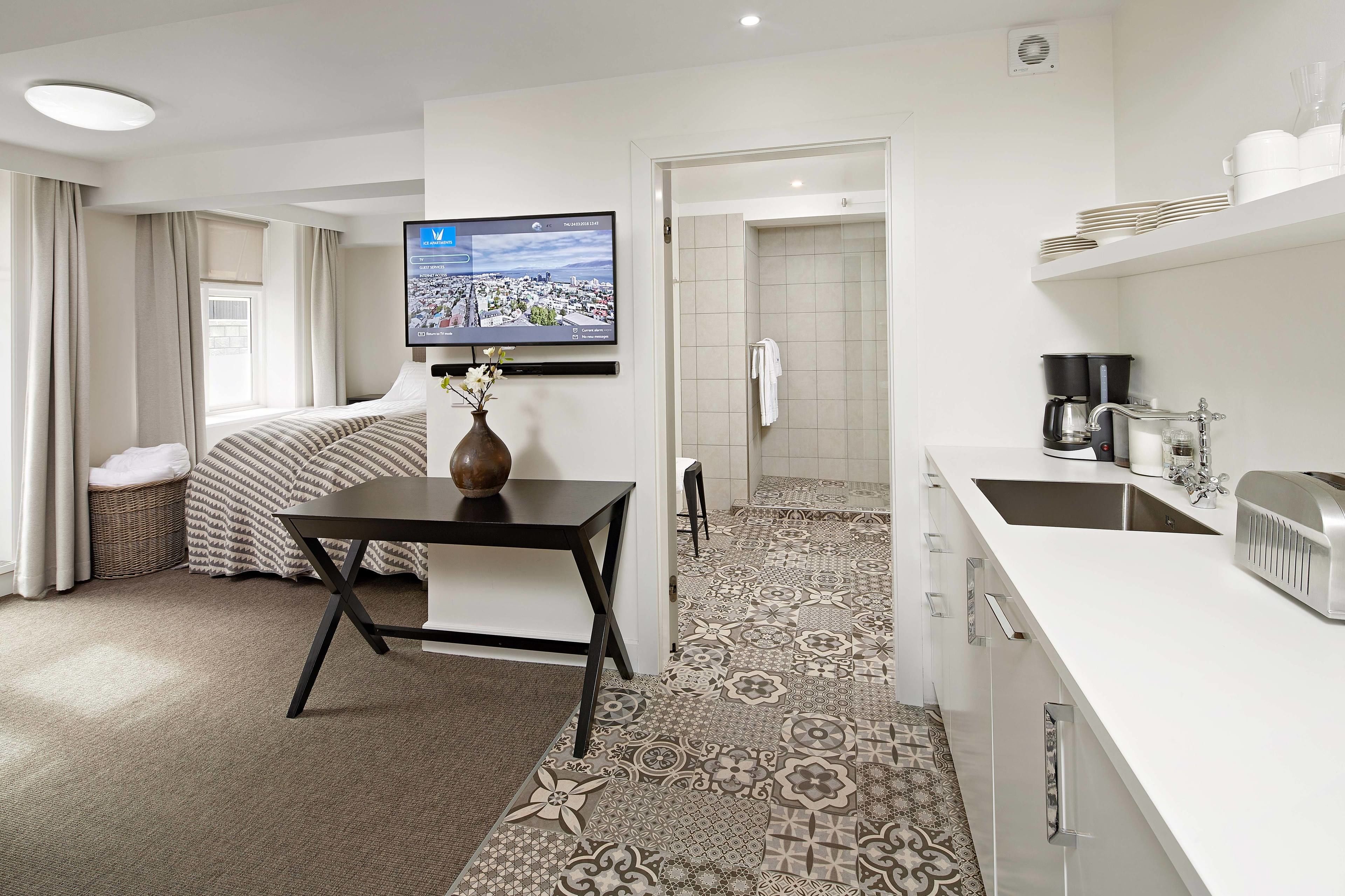 A hotel room with a bed, a TV mounted on the wall, a small table with a vase, a kitchen area with a sink and a toaster, and a patterned floor.