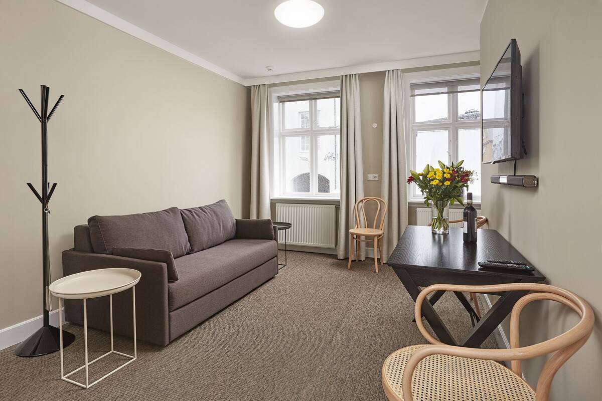 A modern living room with a sofa, a small round table, a dining table with chairs, a floor lamp, and a TV mounted on the wall.