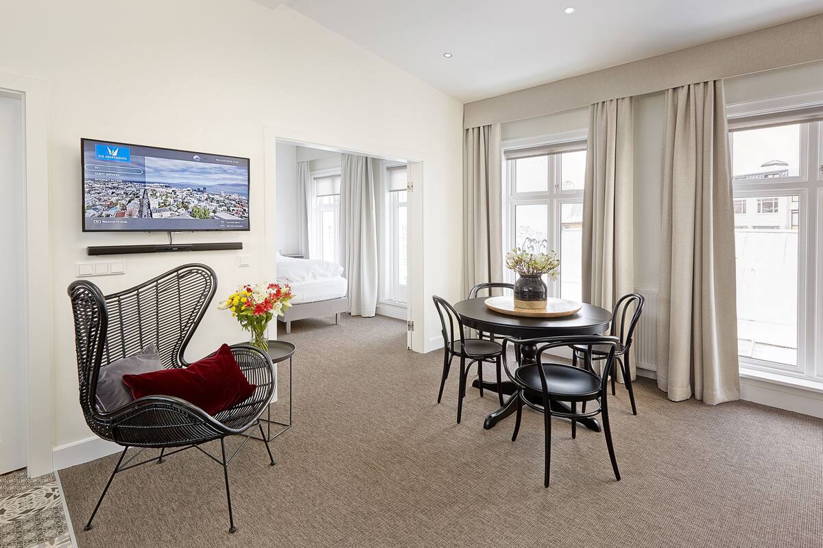 A hotel room with a bed, a TV mounted on the wall, a dining area with a table and chairs, and large windows with curtains.