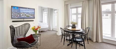 A hotel room with a bed, a TV mounted on the wall, a dining area with a table and chairs, and large windows with curtains.