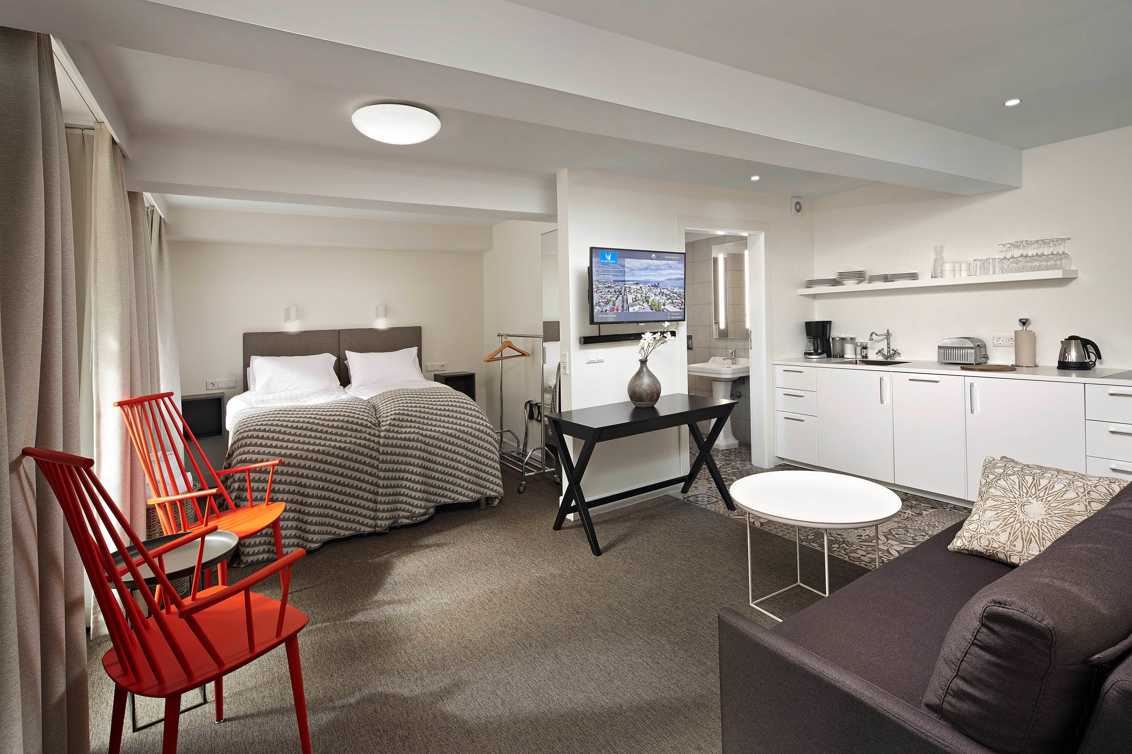 Quarto de hotel compacto com cama, duas cadeiras vermelhas, uma mesinha, um sofá, uma televisão e uma kitchenette.