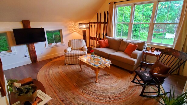 Un salón acogedor con un sofá, un sillón y una mesa de centro, un televisor de pantalla plana montado en la pared, y un ventanal con vistas al exterior.
