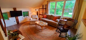 A cozy living room with a sofa, armchair, and coffee table, a flat-screen TV mounted on the wall, and a large window offering a view of greenery outside. - Hobble Inn (Stowe)