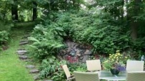 Un jardín con una mesa y sillas, rodeado de exuberante follaje y un muro de piedra.