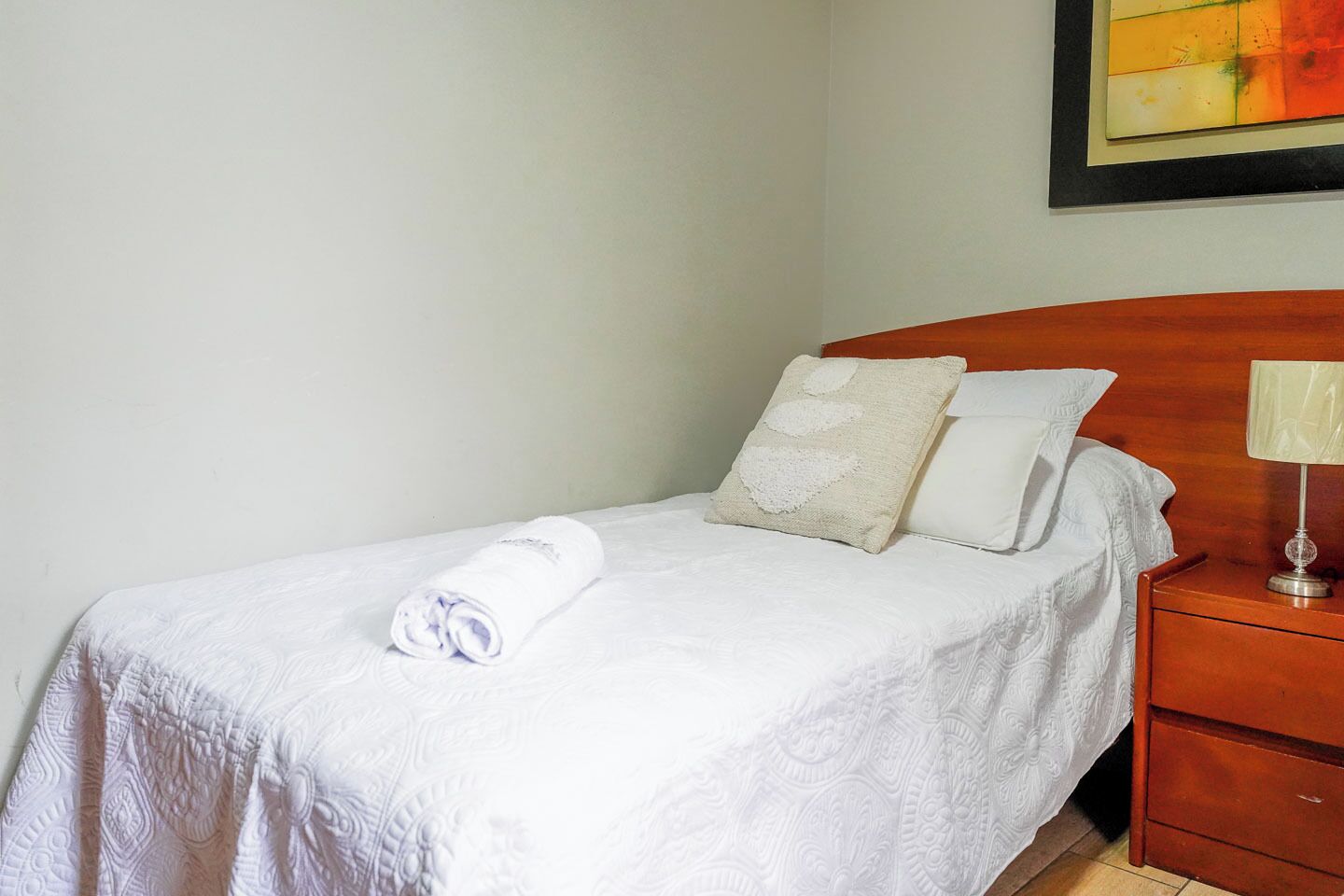 A neatly made bed with a rolled white towel, two pillows, and a wooden nightstand with a lamp.