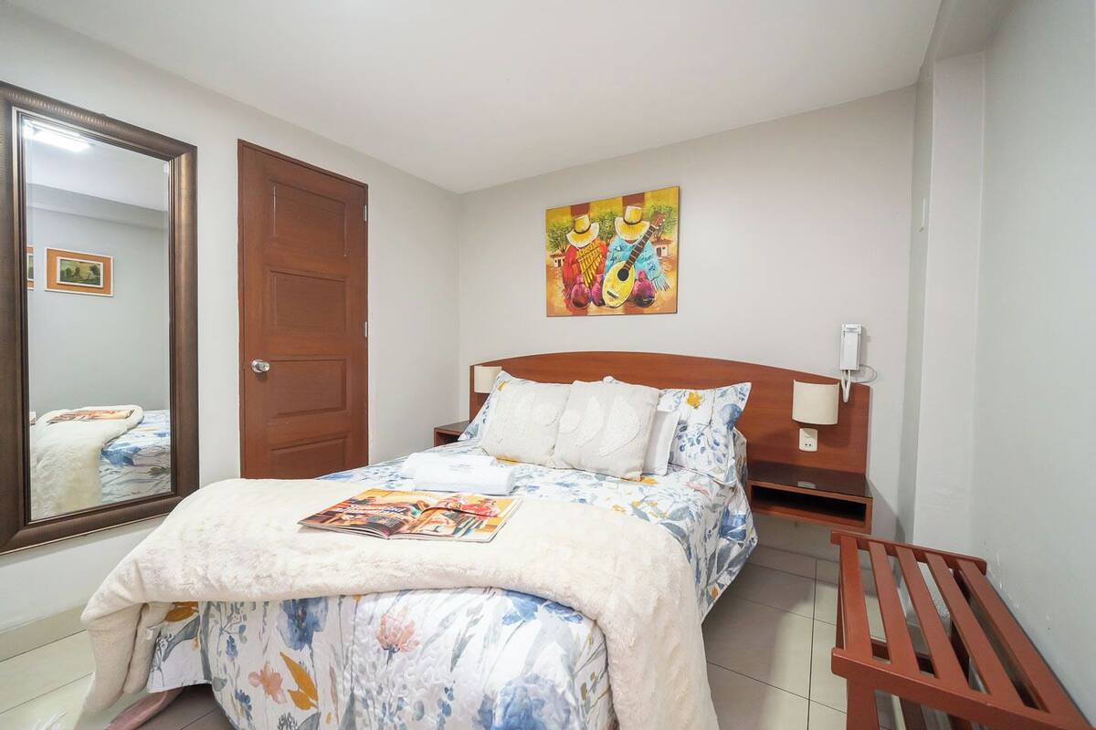 Um quarto com cama, cabeceira de madeira, mesa de cabeceira, abajur, espelho e um quadro na parede.