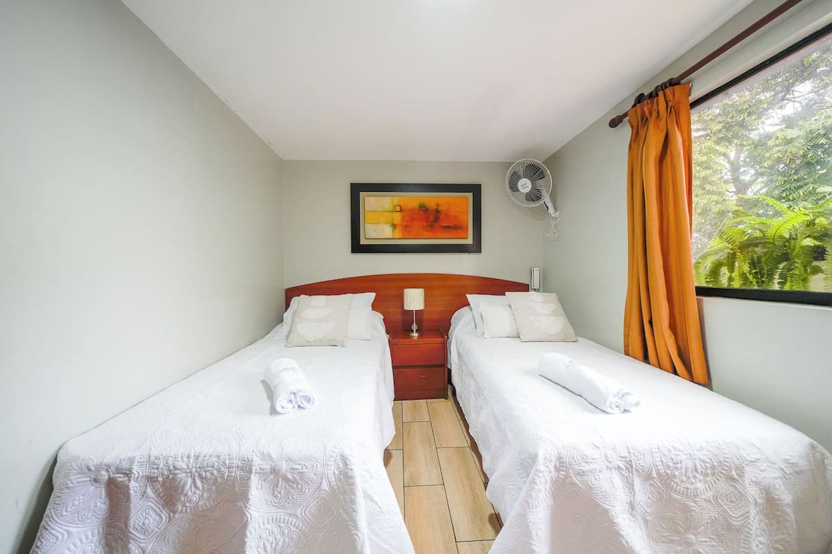 Quarto de hotel com duas camas de solteiro, cabeceira de madeira, um quadro emoldurado, um ventilador de chão e uma janela com cortinas.