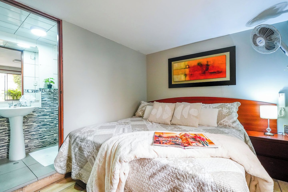A bedroom with a bed, a bedside table with a lamp, a wall-mounted fan, and a painting.