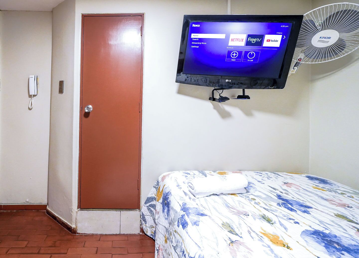 A hotel room with a bed, a television mounted on the wall, a door, and a wall-mounted fan.