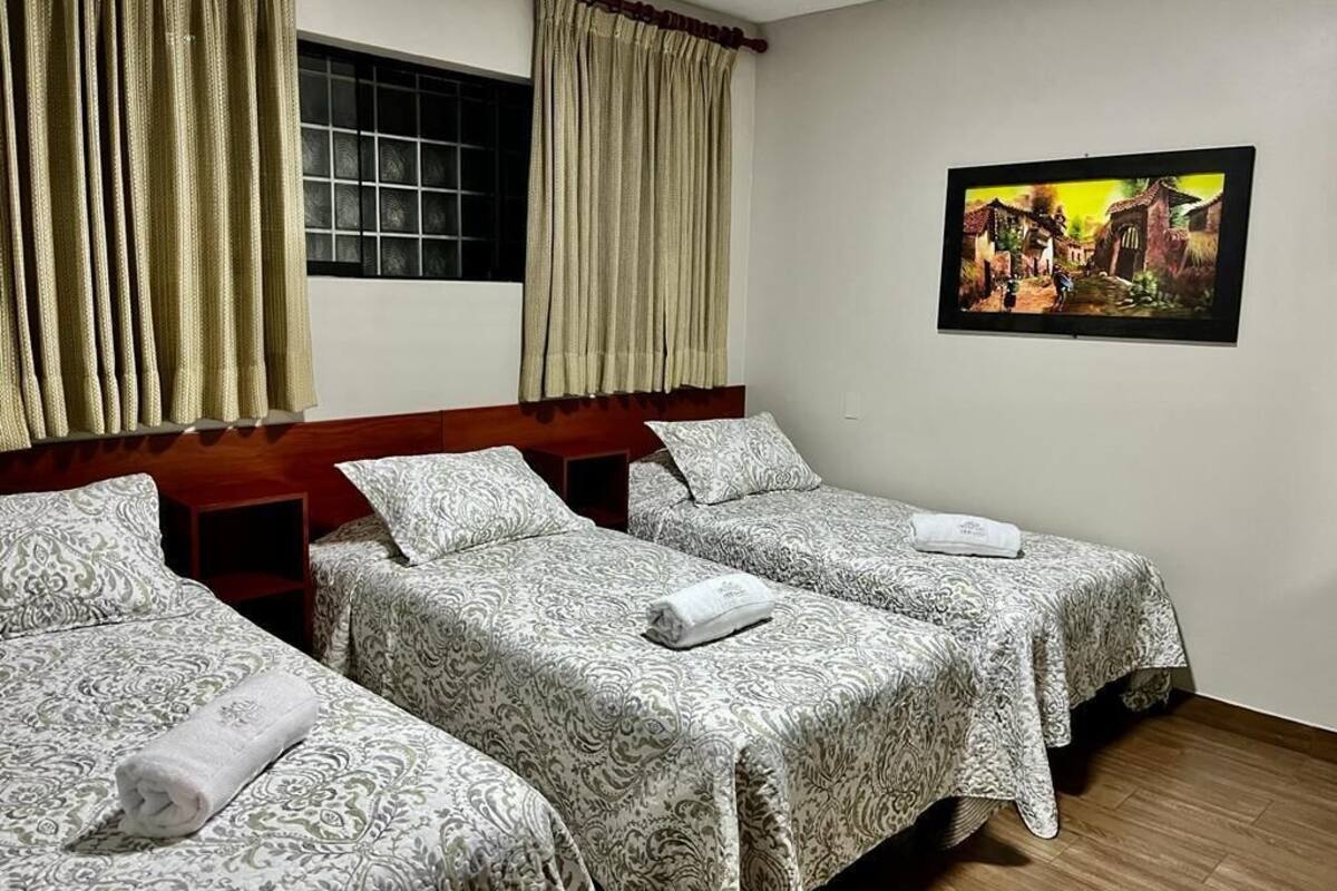 Quarto de hotel com três camas, cortinas bege, janela com vista e um quadro na parede.
