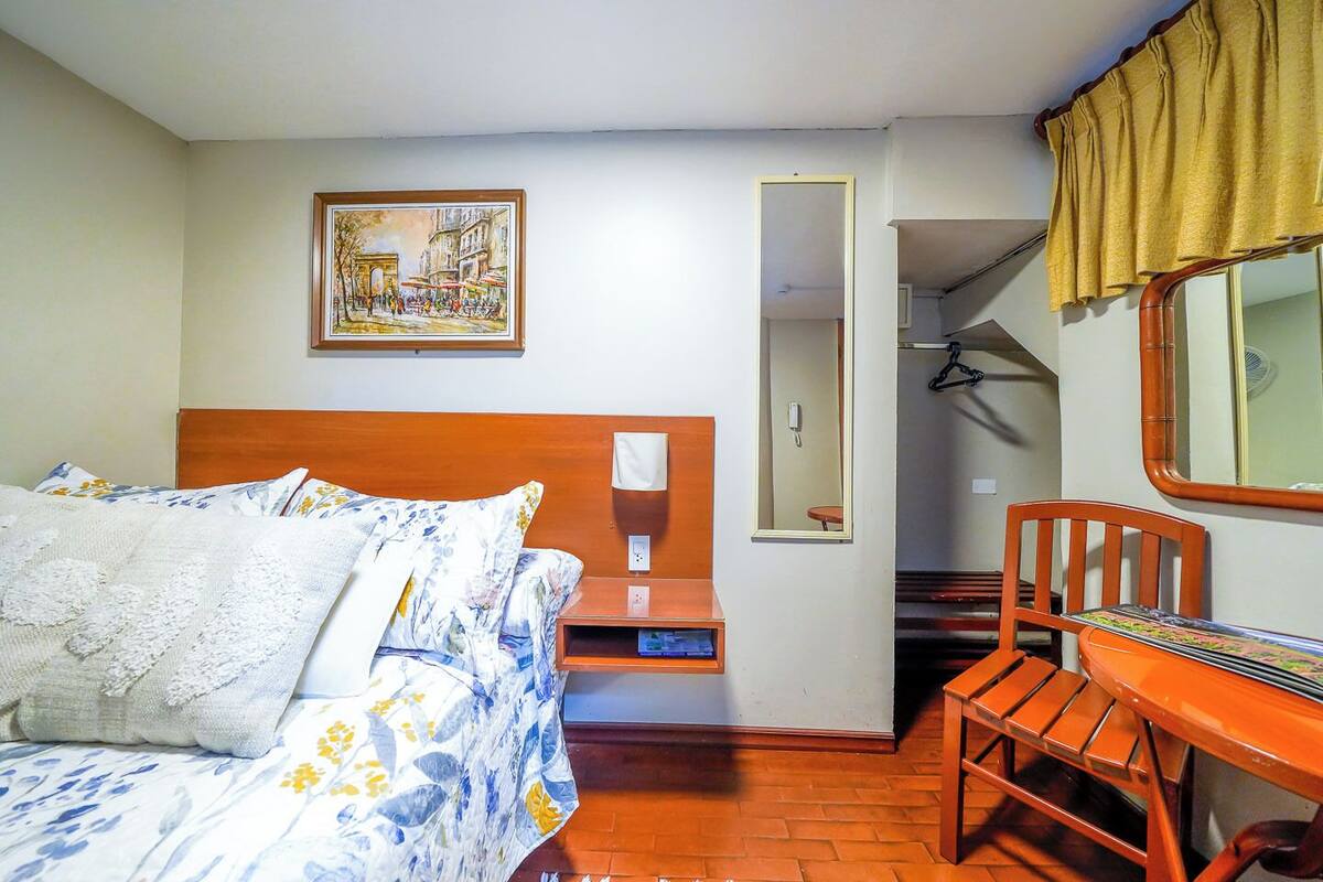 Quarto de hotel com cama, mesa pequena, cadeira, espelho, quadro e escada.