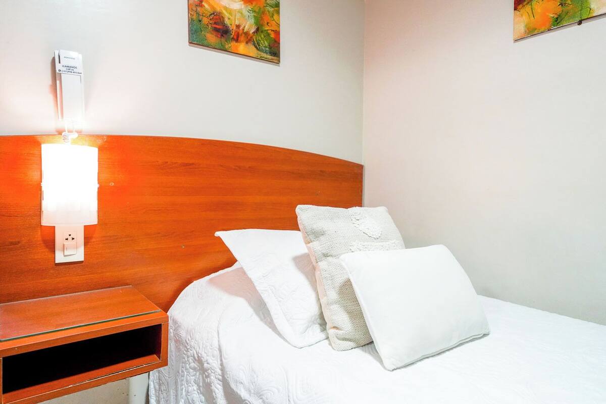 Quarto de hotel com cabeceira de madeira, mesa de cabeceira com abajur e duas almofadas brancas na cama.