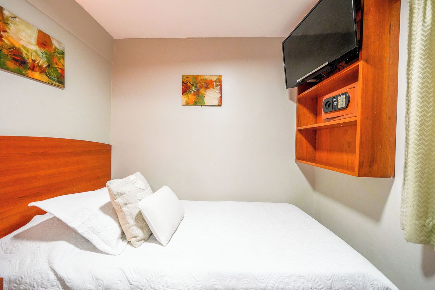 Una cama bien hecha con ropa blanca y almohadas, un cabecero de madera, un televisor montado y dos cuadros en la pared.
